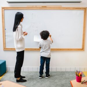 A teacher assists a young boy writing on a whiteboard in an engaging classroom setting.